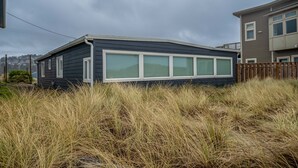 A single-story house with a dark exterior and white-framed windows, surrounded by tall grass. - Beachfront Home, on the Sand and Pet Friendly! (Pacific City)