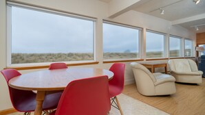A modern interior with large windows, a wooden table, red and white chairs, and a view of the dunes. - Beachfront Home, on the Sand and Pet Friendly! (Pacific City)