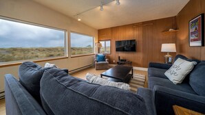 A modern living room with a large window, a flat-screen TV, and a cozy seating area. - Beachfront Home, on the Sand and Pet Friendly! (Pacific City)