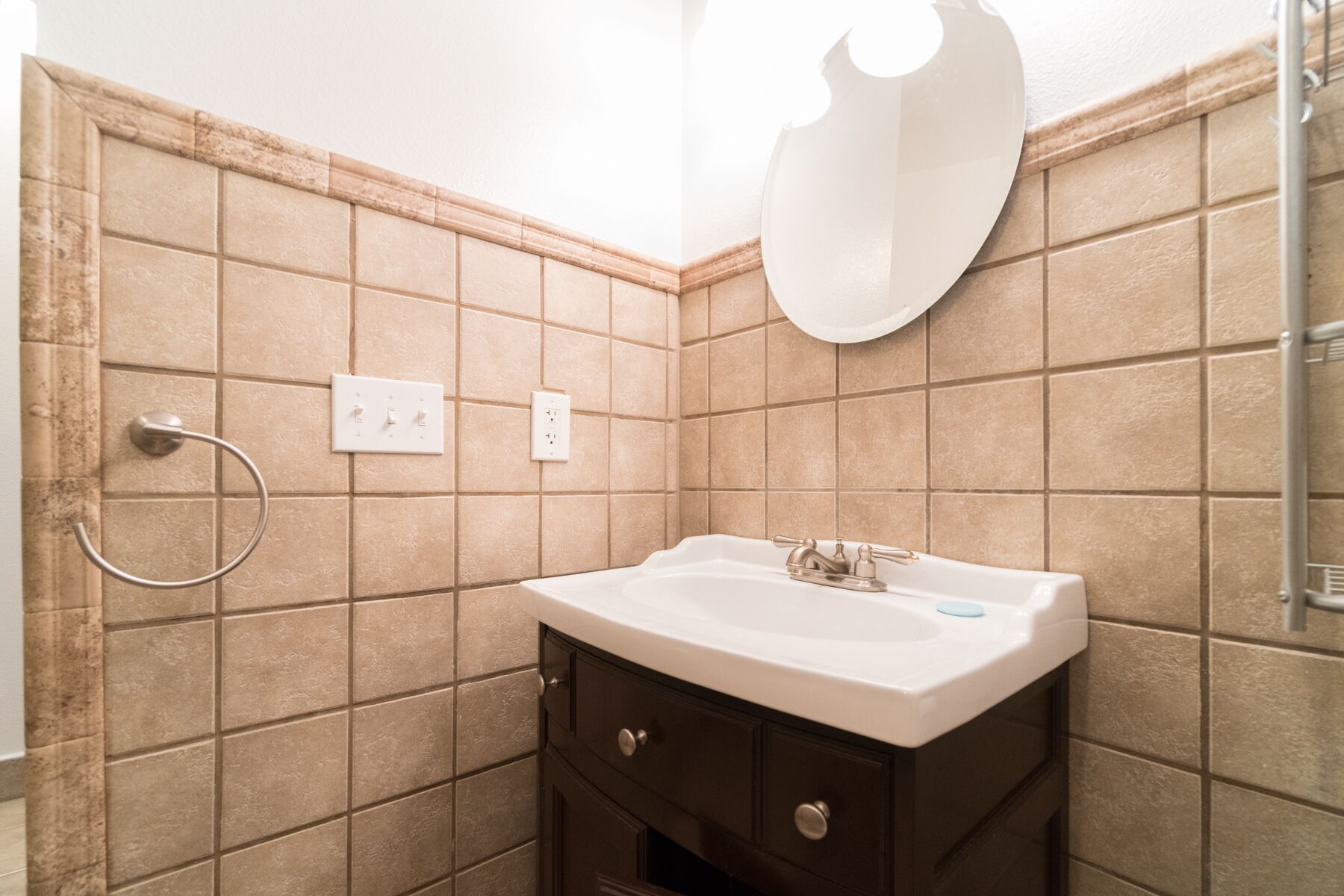 Un baño con baldosas color beige, un lavamanos blanco, un mueble oscuro, un tozalete y un espejo.
