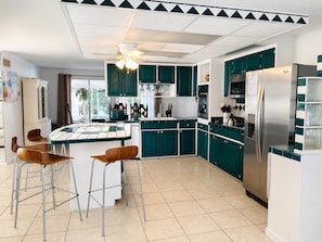 A modern kitchen with teal cabinets, a stainless steel refrigerator, and a dining area with stools.