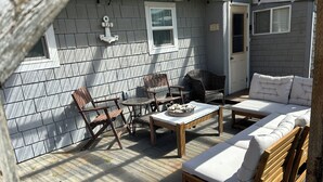 Eine sonnendurchflutete Terrasse mit Holztisch, zwei Stühlen, einem Korbstuhl und einer Couch mit Kissen.