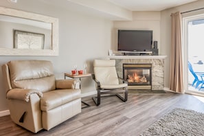 A living room with a leather sofa, a white armchair, a flat-screen TV, a fireplace, and a view through a large window.