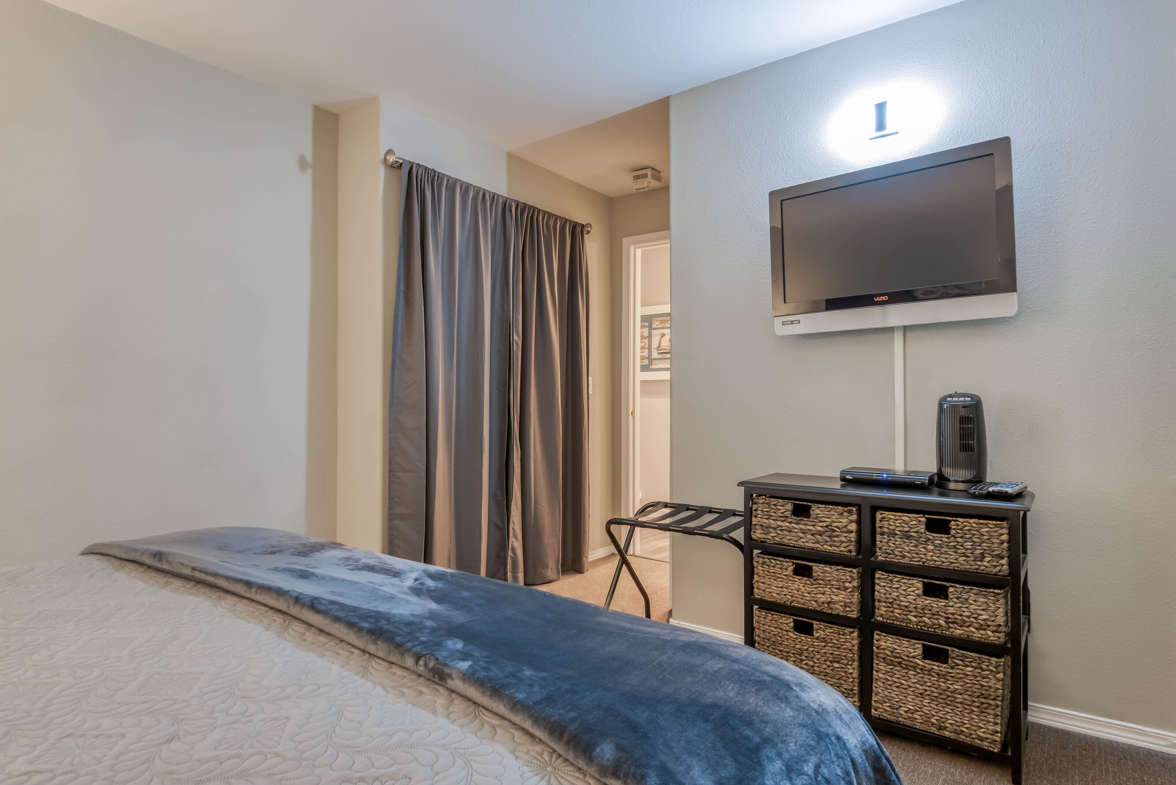 Un dormitorio con una cama, un televisor montado en la pared, una mesita de noche con un altavoz y una puerta que da a otra habitación.