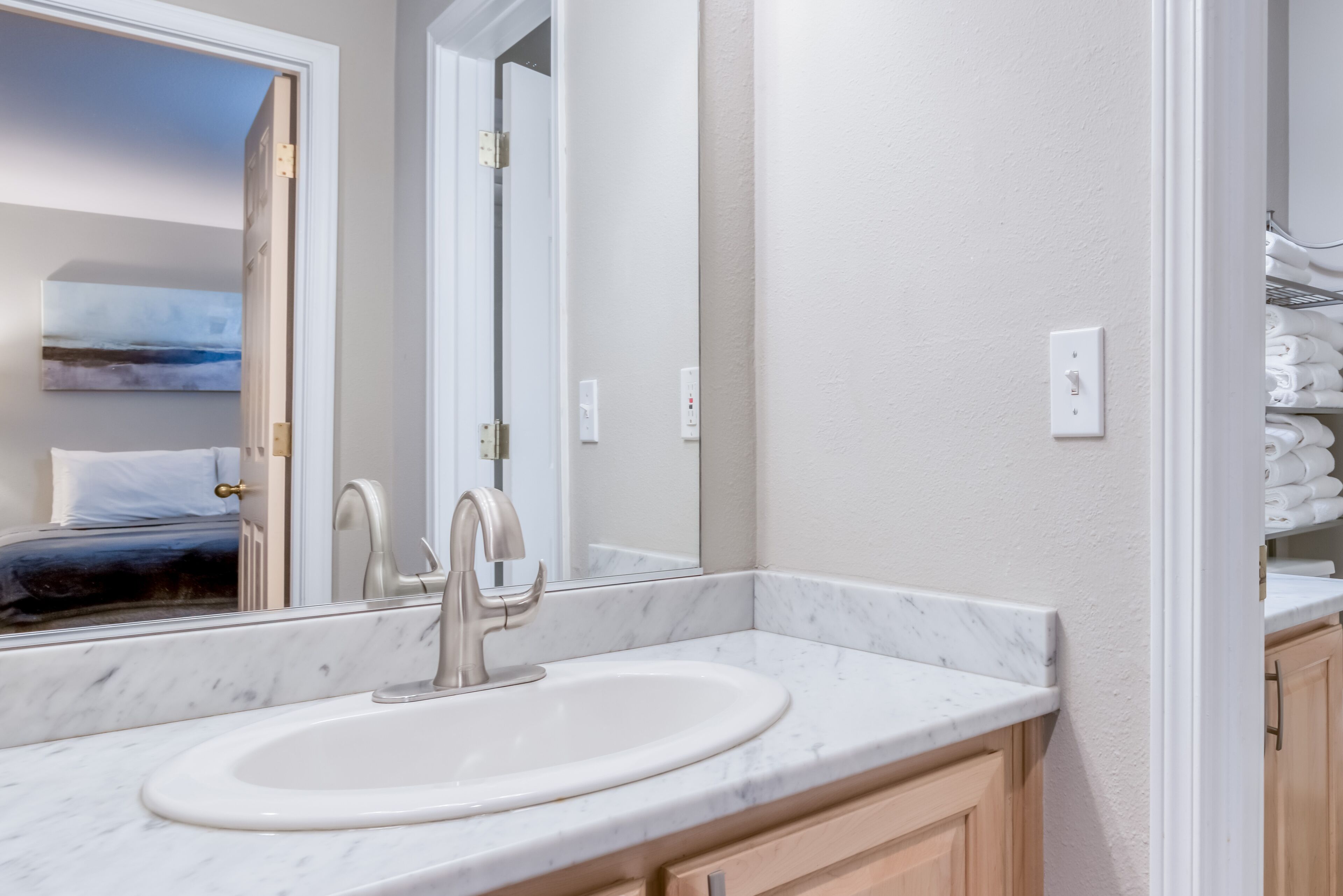 A bathroom with a white sink, a mirror, and a view of a bedroom with a bed and a painting.