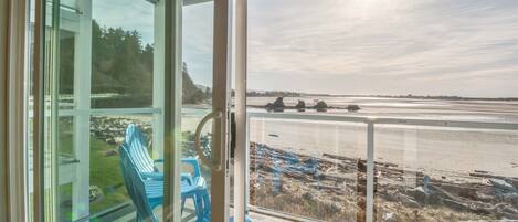 Uitzicht vanuit een kamer met een glazen deur naar een balkon met uitzicht op een strand, met stoelen en een heldere lucht.