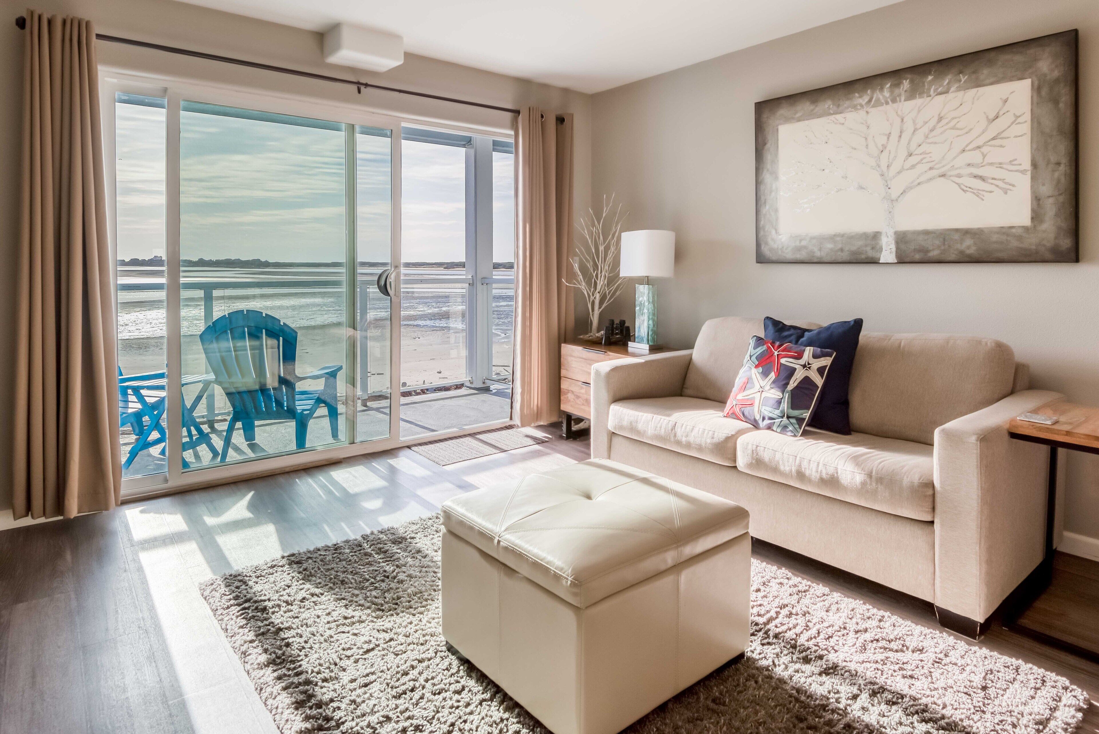 Ein Wohnzimmer mit einer Couch, Hocker und Blick auf den Strand durch große Glasfenster.