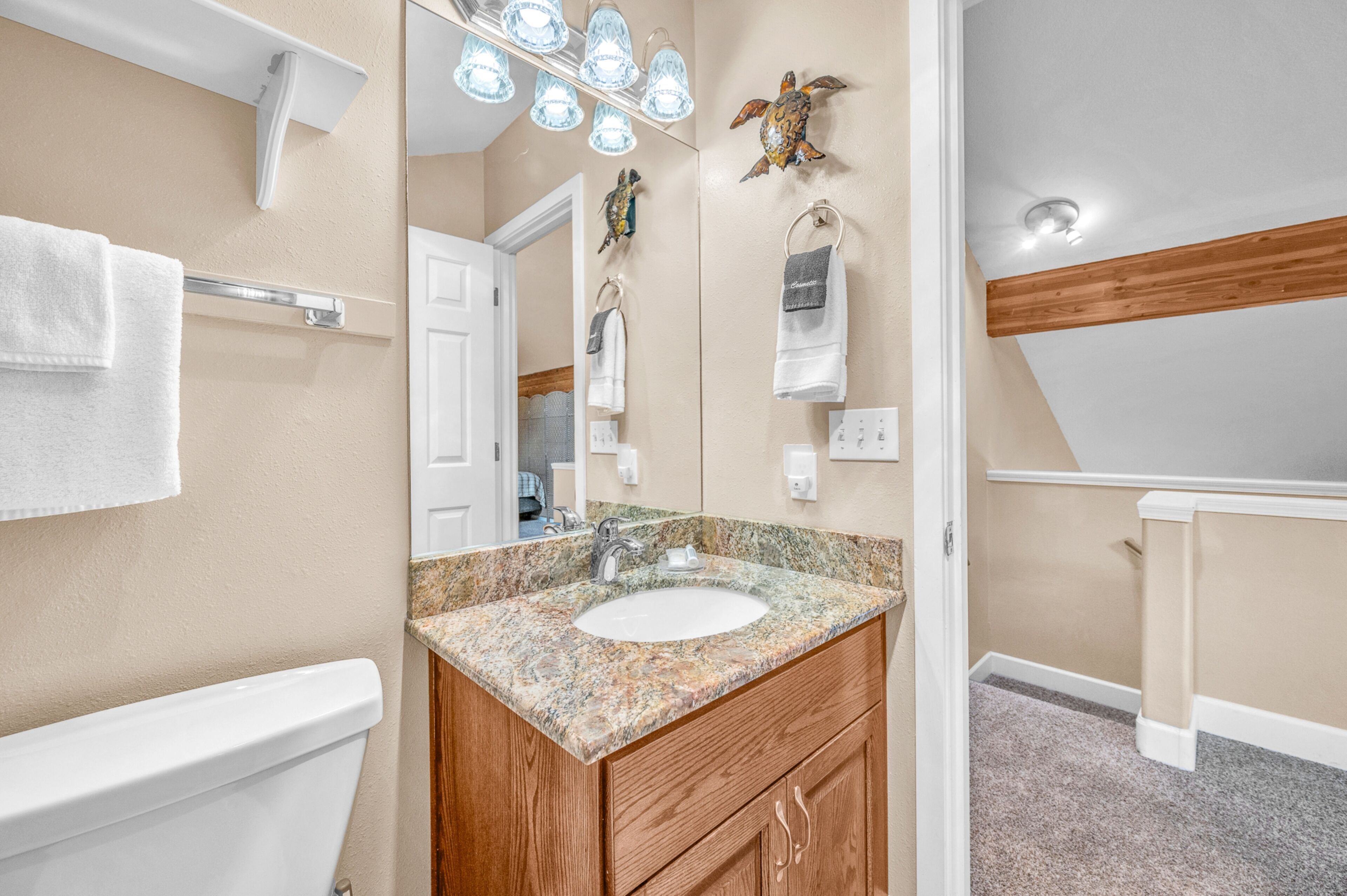 A bathroom with a white toilet, a granite sink, wooden cabinets, a towel rack, a mirror with lights, and a ceiling fan.