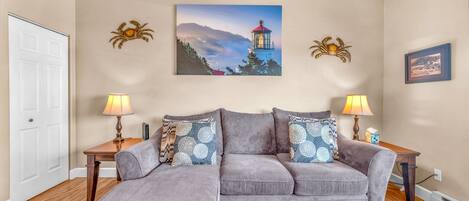 A living room with a sectional sofa, two side tables, a lamp, a painting of a lighthouse, and a door.