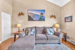 Una sala de estar con un sofá modular, dos mesitas auxiliares, una lámpara, un cuadro de un faro y una puerta.