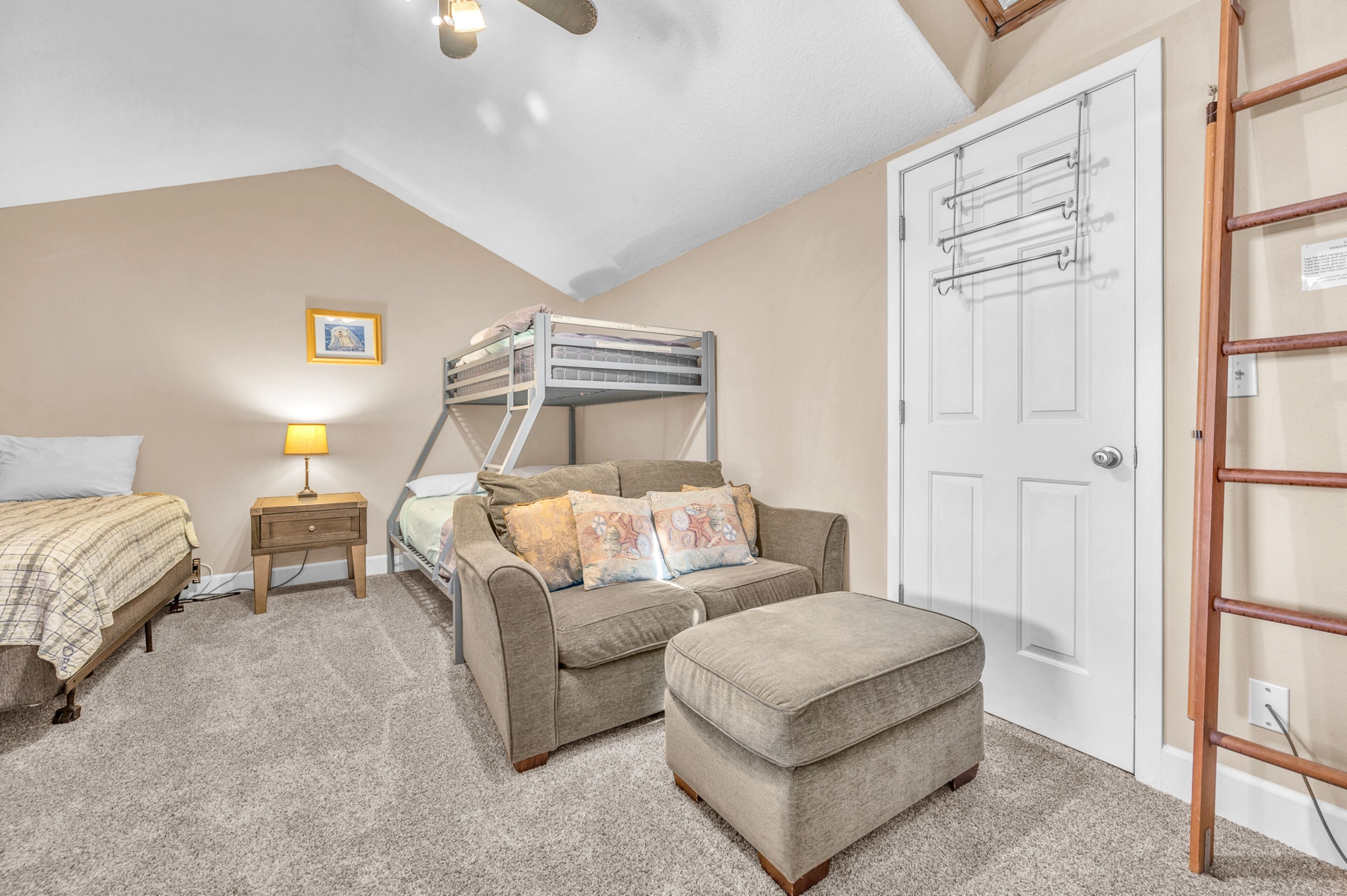 A bedroom with bunk beds, a sofa, an ottoman, a nightstand with a lamp, and a door with a ladder.