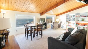 Ein gemütliches Wohnzimmer mit Holzbalkendecke, einem Essbereich mit Stühlen, einer Couch, einem Couchtisch, einem Fernseher und einem Bücherregal voller Bücher und Spielzeug.