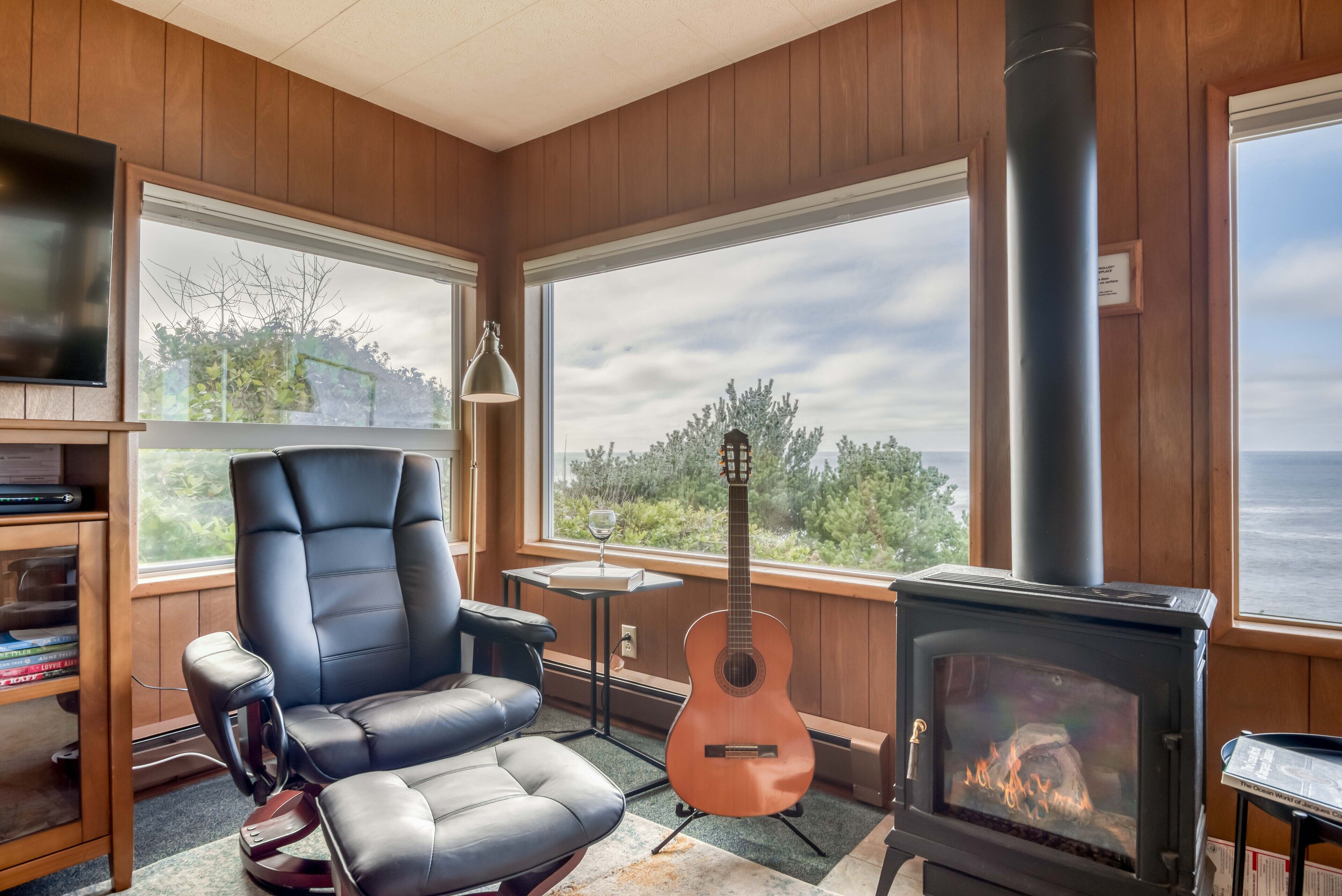 A cozy room with a fireplace, a comfortable chair, a guitar, and a view of the outdoors.