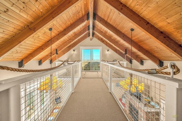 A high-ceilinged attic room with a rope railing, a view through a sliding door, and a carpeted floor. - Beautiful home with ocean views, easy beach access and wheelchair accessible (Lincoln City)