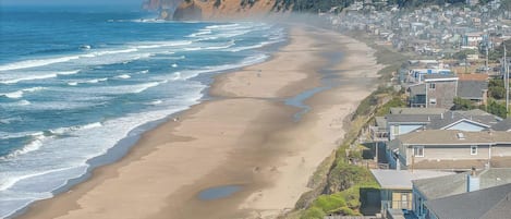A coastal town with a sandy beach, houses along the shore, and a forested hill in the background.
