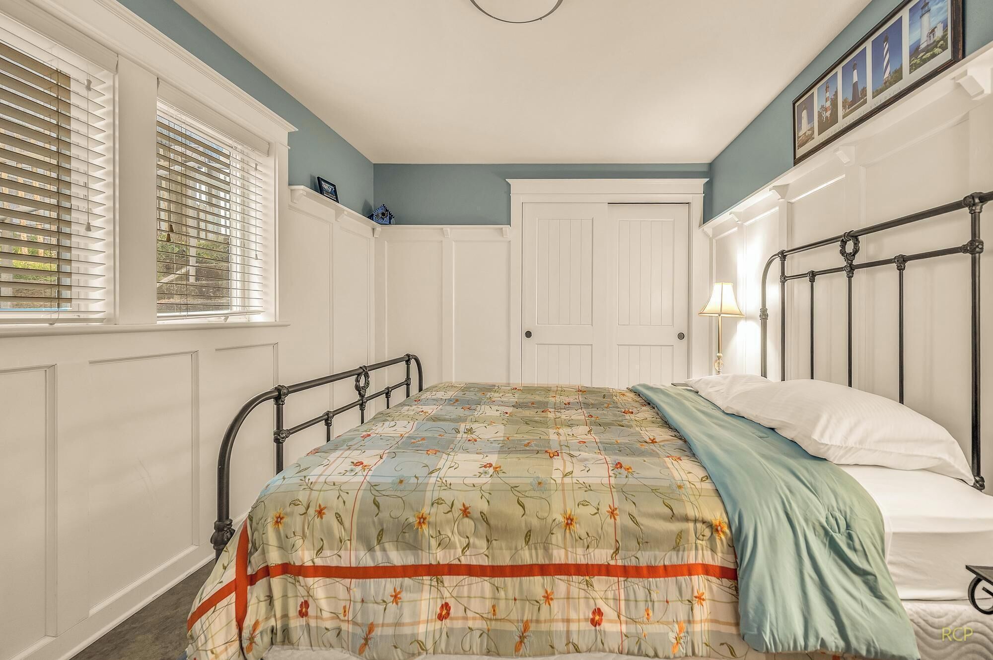 Un dormitorio con cama, ventana con persianas, una puerta y una pared con cuadros.