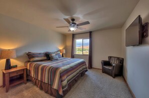 A bedroom with a bed, bedside lamps, a chair, a ceiling fan, and a window with a view. - Southgate 4 by MoabCondos4Rent (Moab)