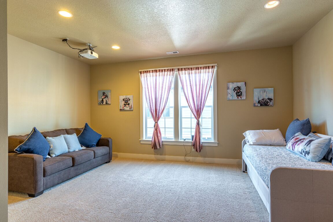 A room with beige walls, two windows with pink curtains, a brown sofa with blue cushions, a white sofa with patterned cushions, and a ceiling projector.