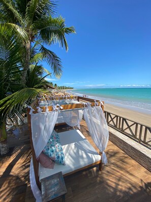 Una cama frente a la playa, con vista al océano, una terraza de madera y palmeras.