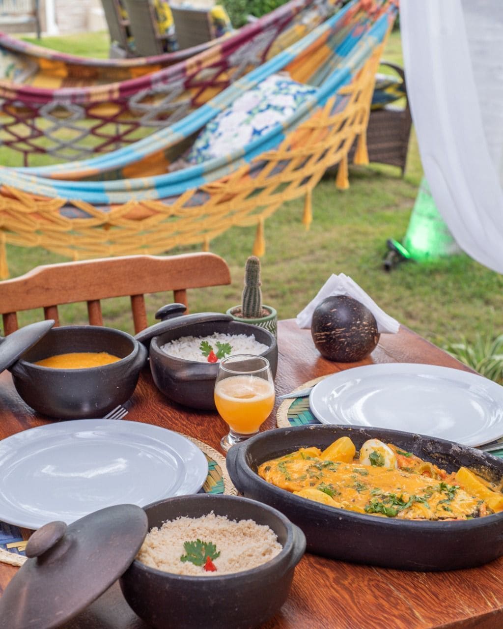 Mesa de comedor al aire libre con diversos platos, una hamaca y un entorno rústico.