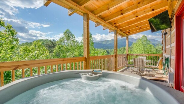 A hot tub with a control panel, surrounded by a wooden deck and a red door, overlooking a mountainous landscape. - Mount LeConte Views, Genuine Log Cabin, Hot Tub, Easy Access Near Town, Fire Pit (Gatlinburg)