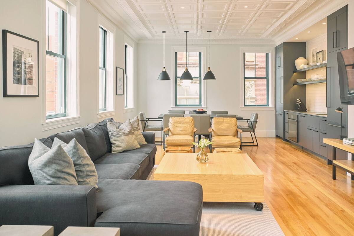 A modern living room with a sofa, coffee table, and chairs. There is a kitchen area with cabinets and a refrigerator.