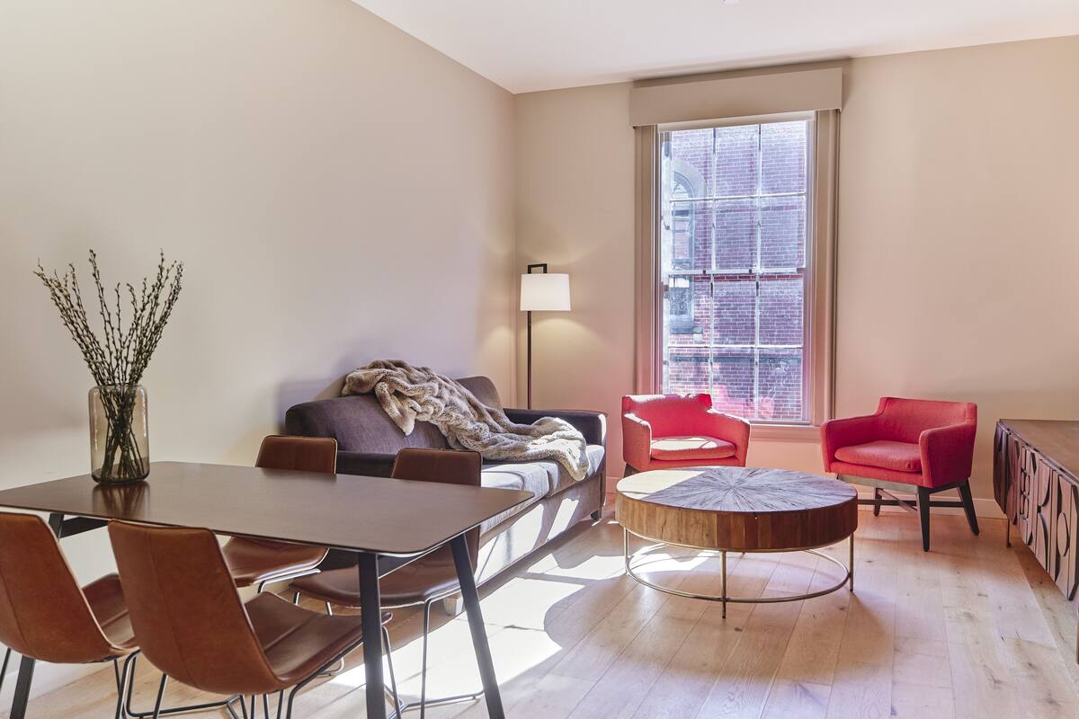 A modern living room with a dining table, two sofas, a red chair, a lamp, and a large window with a view of a brick building.