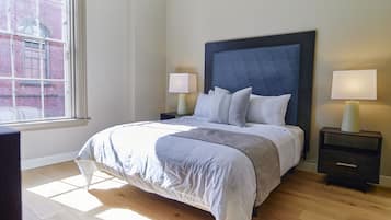 A neatly made bed with a dark headboard, two bedside tables with lamps, and a large window with a view of a brick building.