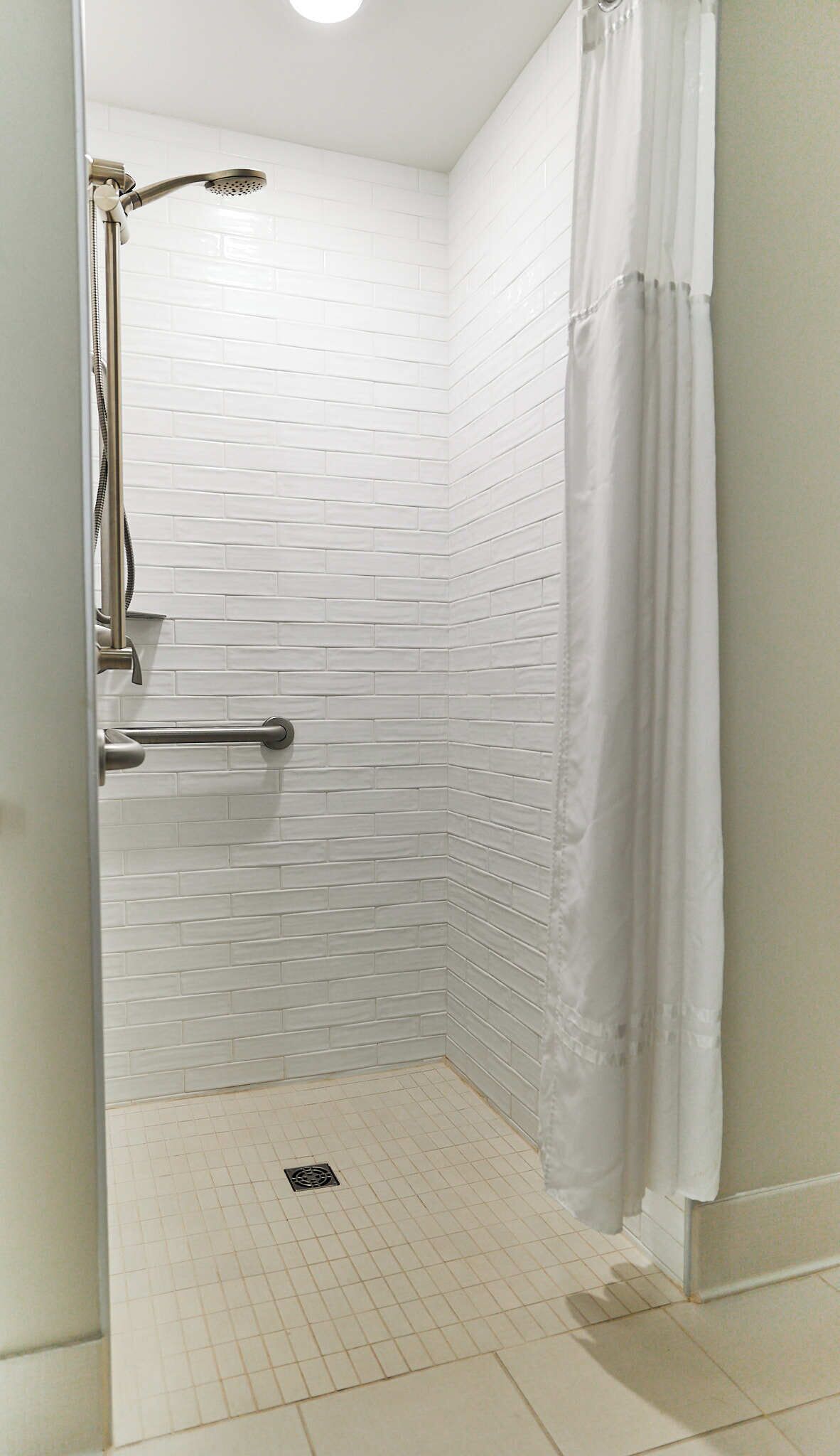 A shower with white tiled walls, a handheld showerhead, a grab bar, and a white curtain.