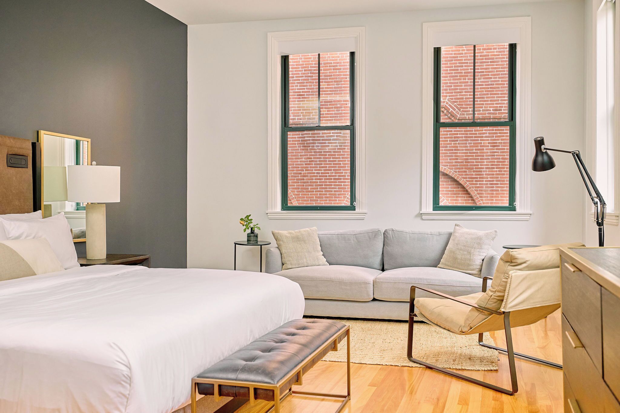 A modern bedroom with a large bed, a sofa, a desk, and two windows with brick walls.