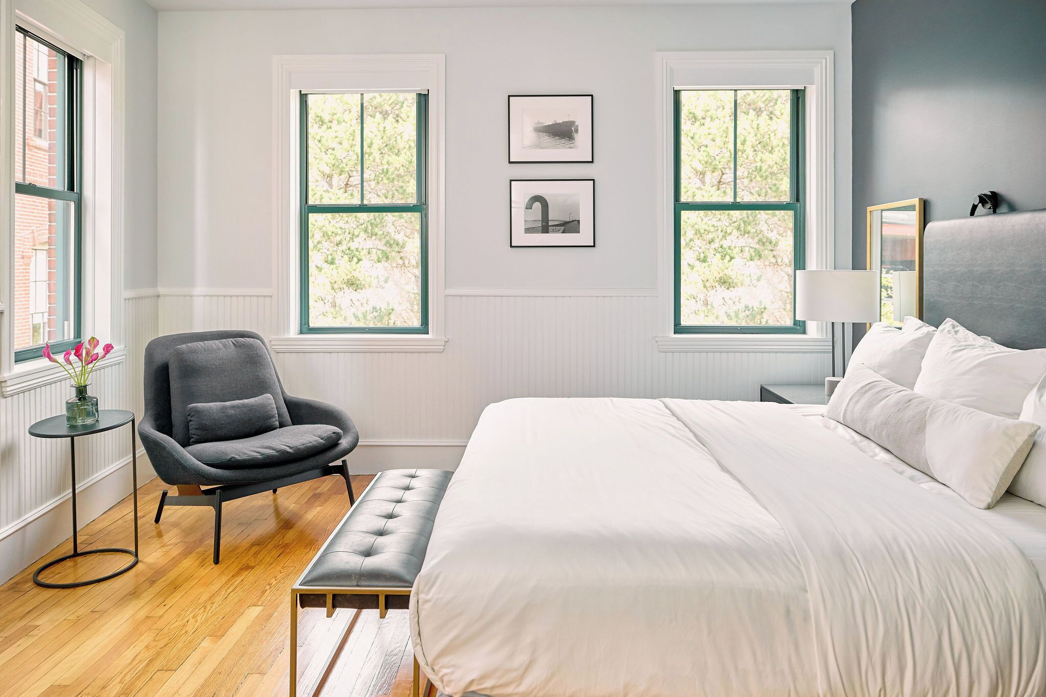 A bedroom with a bed, a chair, a small table, two windows, and wall-mounted artwork.