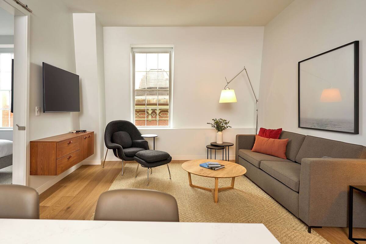 A modern living room with a sofa, armchair, coffee table, TV, and a window with a view.