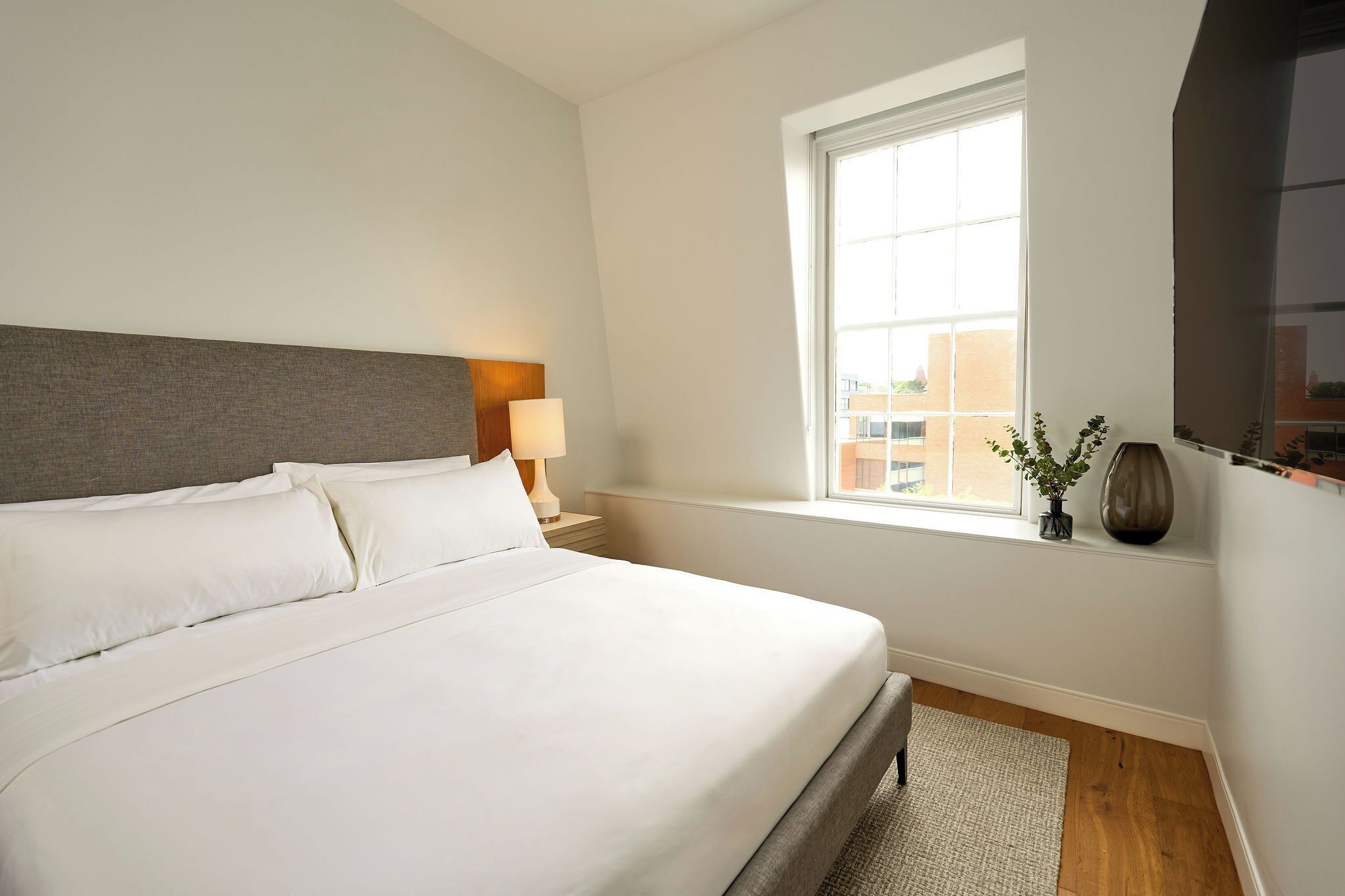 A neatly made bed with white linens, a bedside lamp, a window with a view of buildings, and a vase with plants on the windowsill.