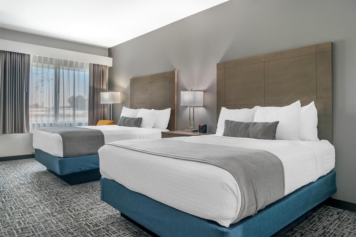 A hotel room with two beds, a large window with curtains, a nightstand with a lamp, and a view of the outdoors.