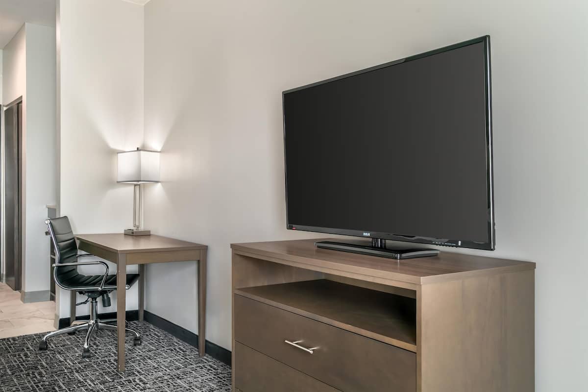 A hotel room with a flat-screen TV mounted on the wall, a desk with a chair, and a lamp.