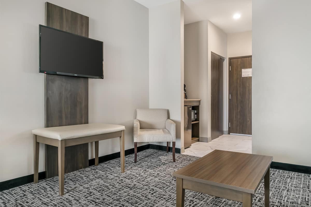 A modern hotel room with a flat-screen TV mounted on the wall, a wooden bench, a patterned rug, and a wooden table.