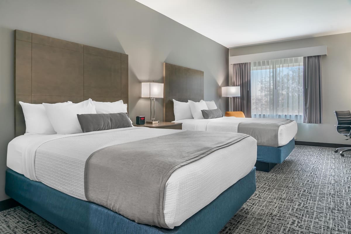 A hotel room with two beds, a large window with curtains, a flat-screen TV, and a desk with a lamp.