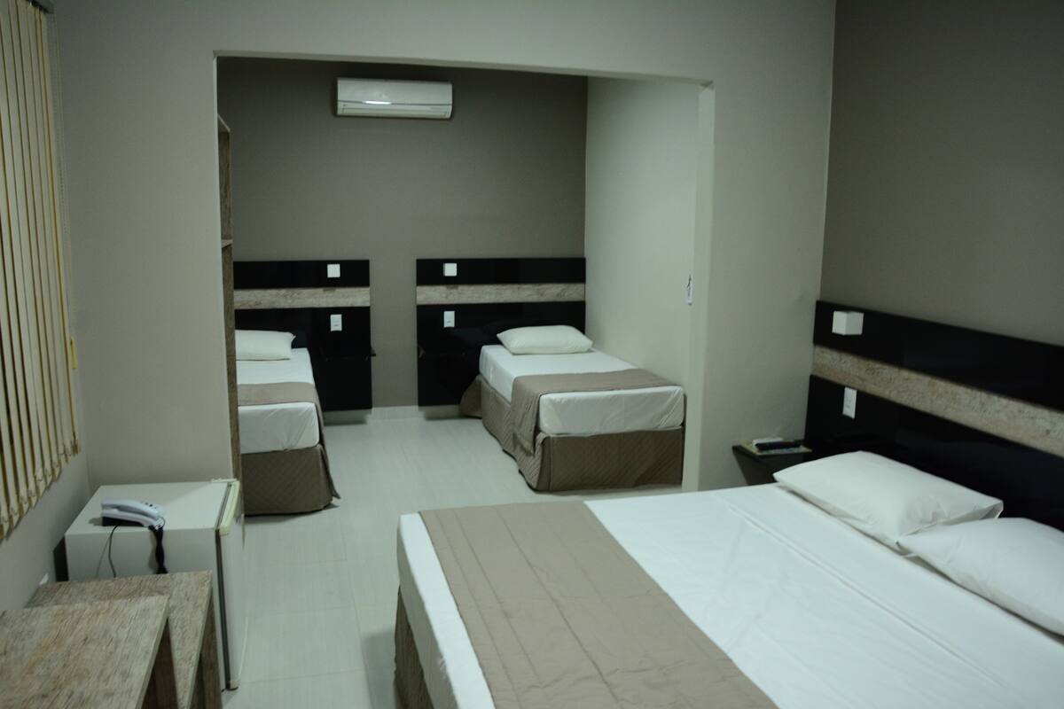A hotel room with two single beds, a larger bed, a desk, a chair, and a small table.