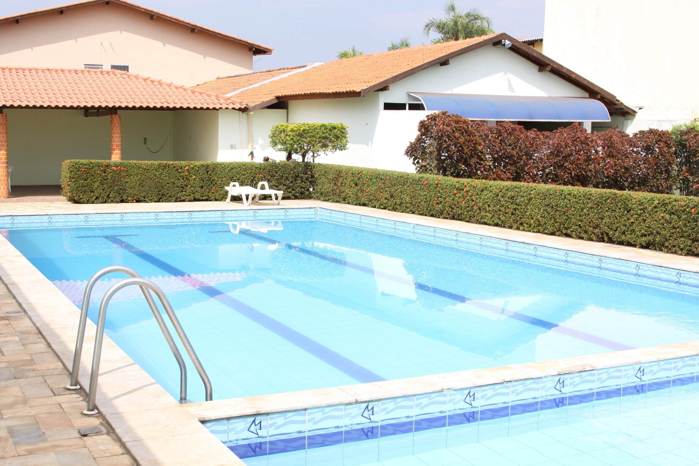 A swimming pool with a tiled edge, a metal ladder, and a white building with a tiled roof in the background. - Riviera Pantanal Hotel (Caceres)