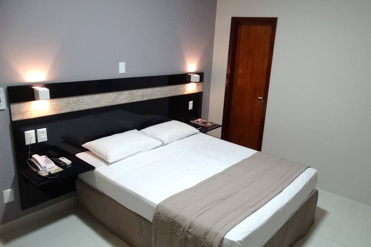 A neatly made bed with white linens and a beige blanket, a bedside table with a phone and remote control, and a wall-mounted light fixture.