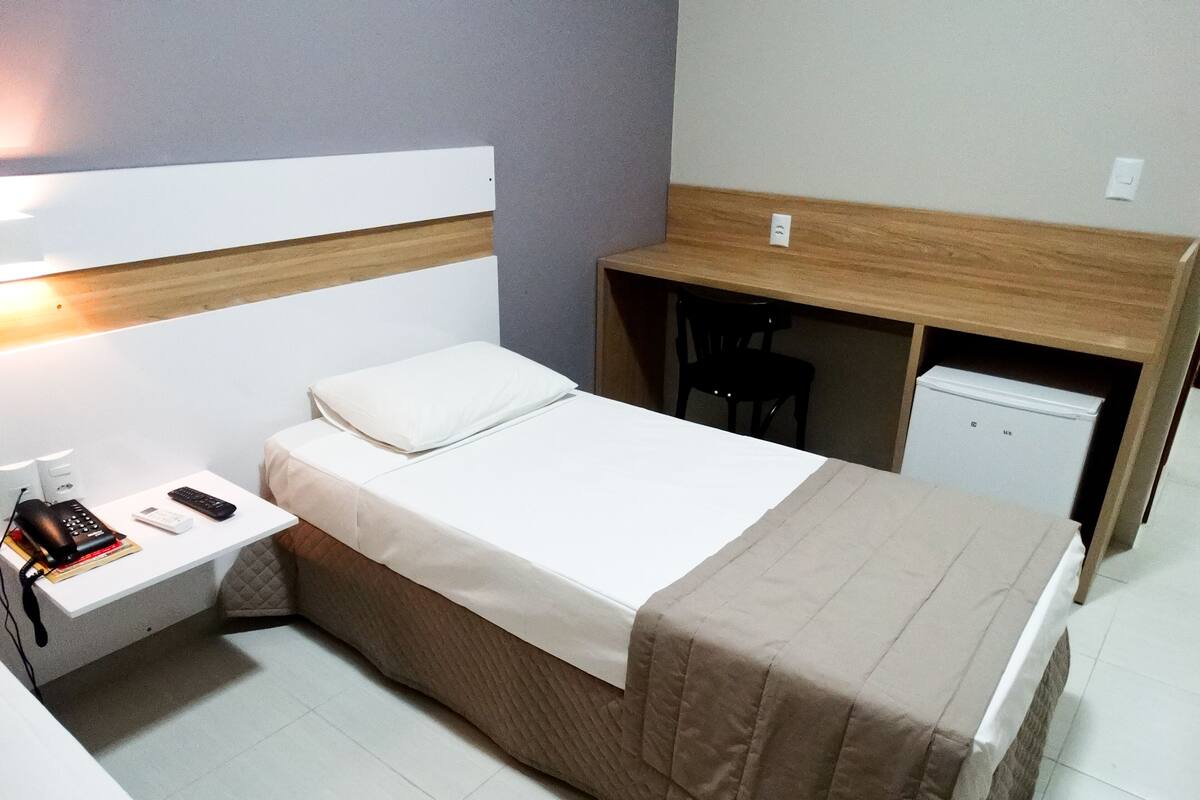 A compact hotel room with a single bed, a bedside table with a phone and books, a small desk with a chair, and a mini-fridge.