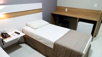 A compact hotel room with a single bed, a bedside table with a phone and books, a small desk with a chair, and a mini-fridge.