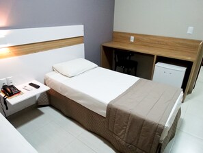 Habitación de hotel compacta con una cama individual, una mesita de noche con un teléfono y libros, un escritorio pequeño con una silla y un mini refrigerador.