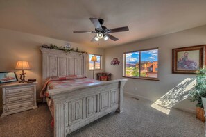 Une chambre à coucher avec un lit, un ventilateur de plafond, une commode, une lampe, une fenêtre donnant sur un beau paysage et un tableau encadré accroché au mur.