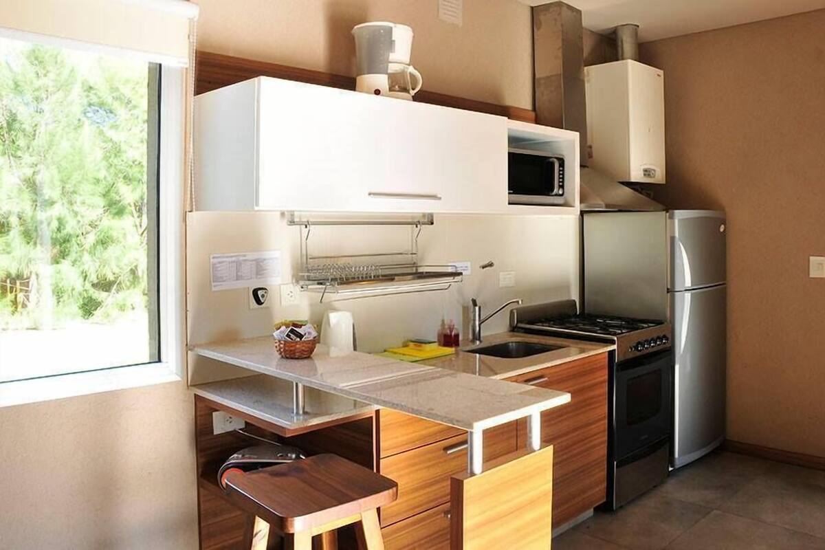 Cocina compacta con microondas, estufa y refrigerador. Se aprecia una ventana con vista a árboles.
