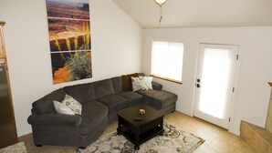 A living room with a gray sectional sofa, a coffee table, a ceiling fan, and a large landscape painting on the wall. - Desert Winds 19 by MoabCondos4Rent (Moab)
