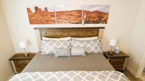 A neatly made bed with patterned bedding, two bedside lamps, and a large landscape painting above the headboard. - Desert Winds 19 by MoabCondos4Rent (Moab)