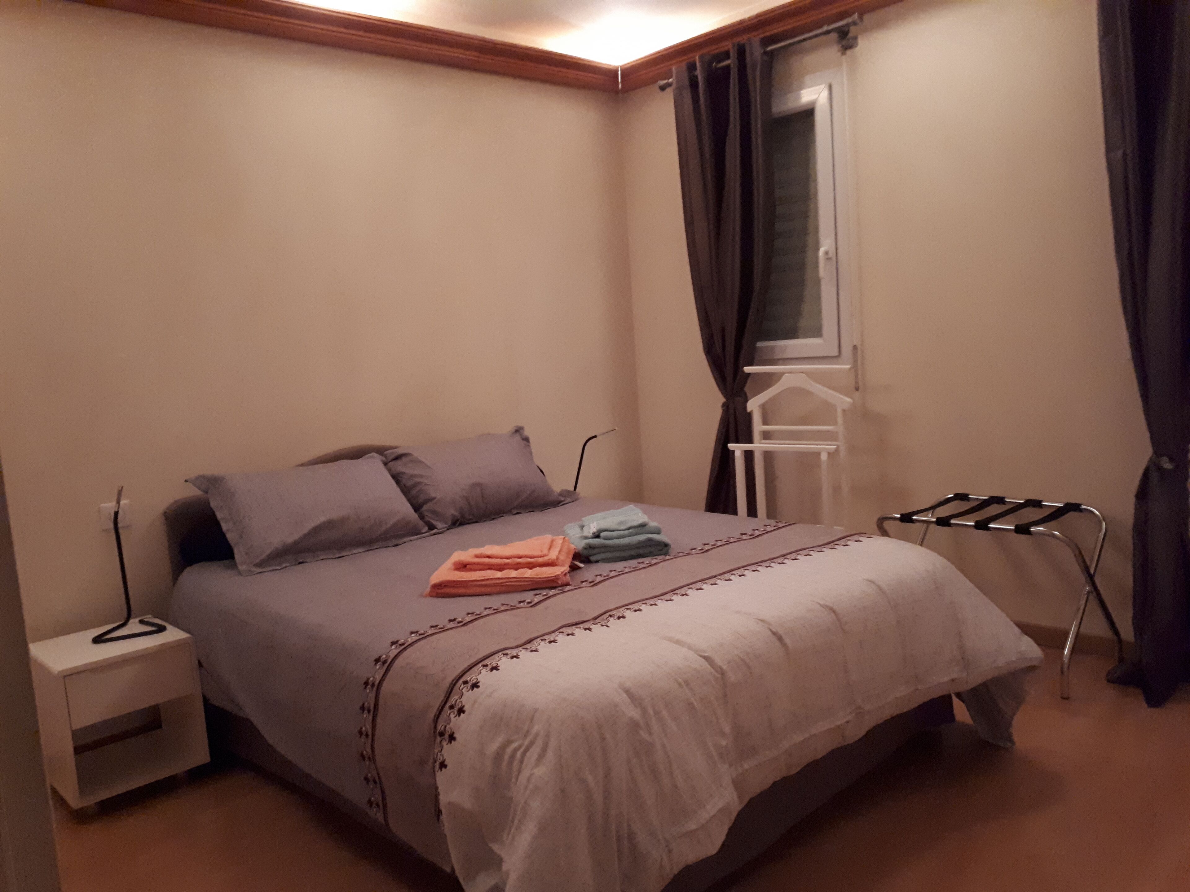 A bedroom with a bed, a nightstand, a window with curtains, and a drying rack.