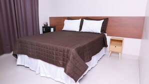 A neatly made bed with a brown quilt and white pillows, a wooden nightstand with a drawer, and a dark-colored cabinet against a white wall. - Hotel Borges (Quirinopolis)