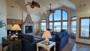 A spacious living room with a fireplace, large windows offering a view of the beach, and ceiling fans. - Oceanfront, Heated Pool, Elevator, Book today! (Corolla)